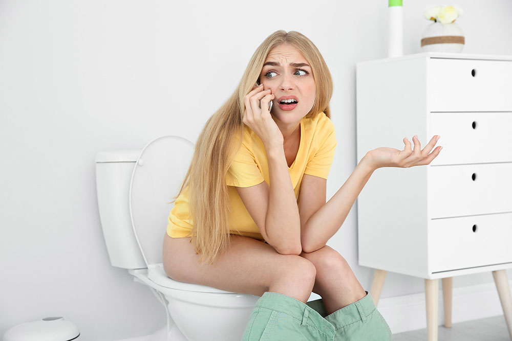 Young woman with mobile phone sitting on toilet bowl at home.
